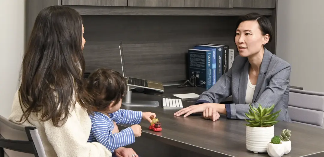 Dr. Esther Chin talks to a parent of a toddler in her office about the neuropsychological testing that is about to transpire in the NeuroMInd office in Schaumburg, IL. The toddler is on his mom's lap playing with a truck.